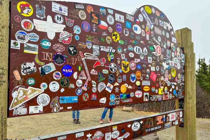 Signboard covered with various colorful stickers and people standing in the background.
