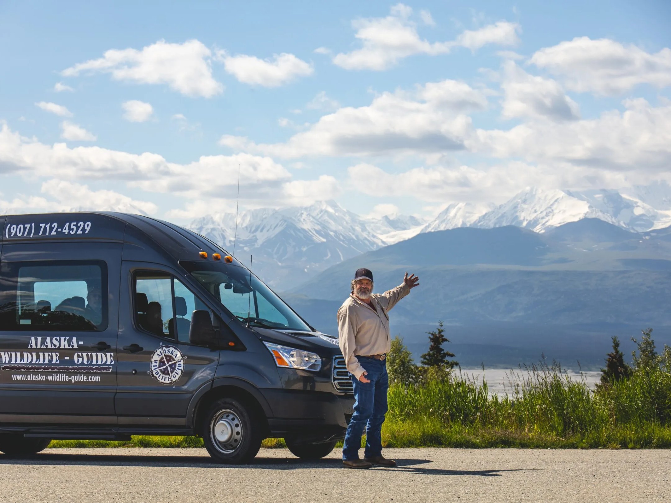 Man standing by Alaska Wild Lights tour van with snow-capped mountains and blue sky in the background