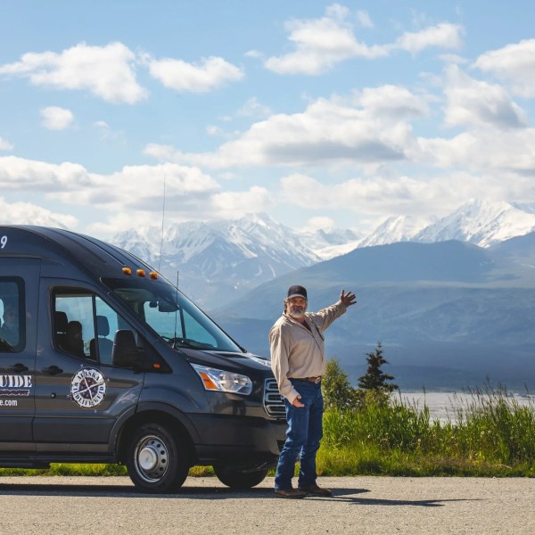 Man standing by Alaska Wild Lights tour van with snow-capped mountains and blue sky in the background