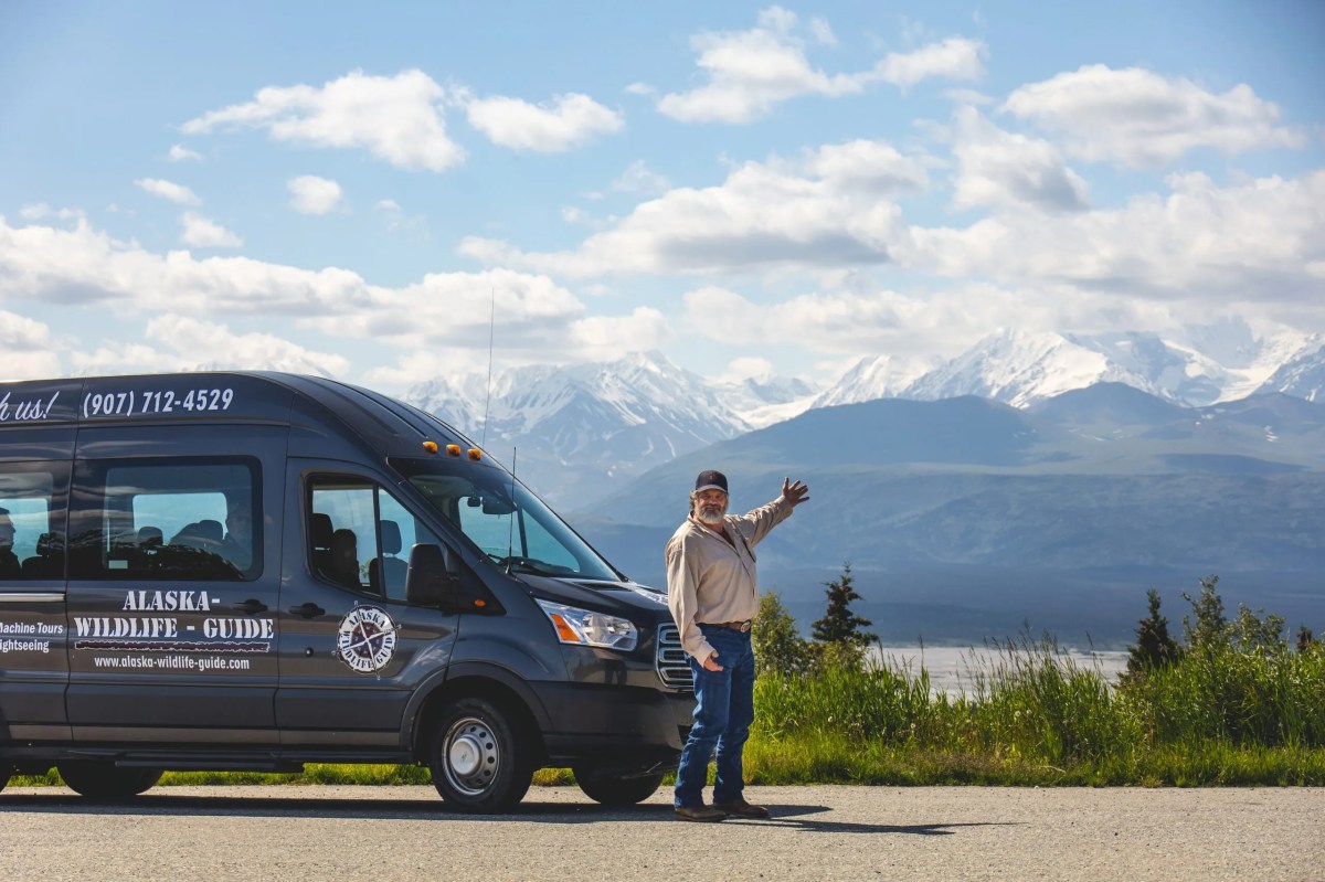 Man standing by Alaska Wild Lights tour van with snow-capped mountains and blue sky in the background