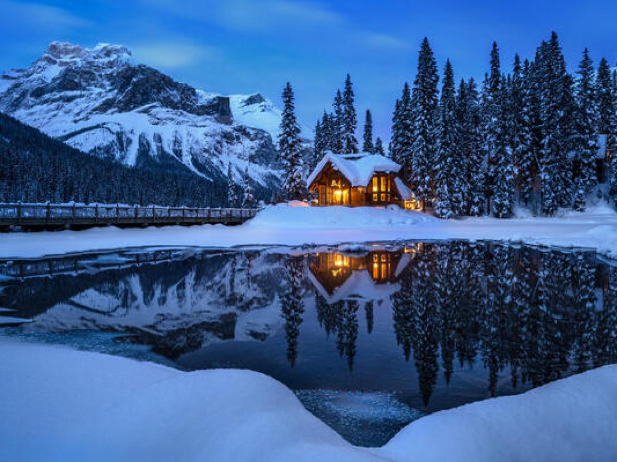 Snowy cabin at dusk by a reflective lake with mountains and pine trees.