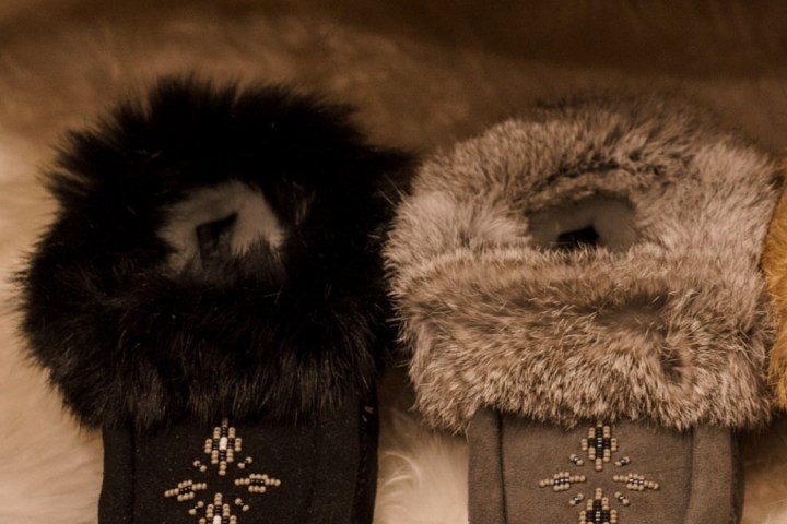 Two pairs of fur-lined moccasins with bead patterns, one black and one gray, on a fluffy white rug.
