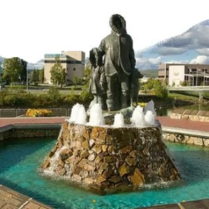 Stone fountain with statues in a park, surrounded by trees and buildings under a cloudy sky.