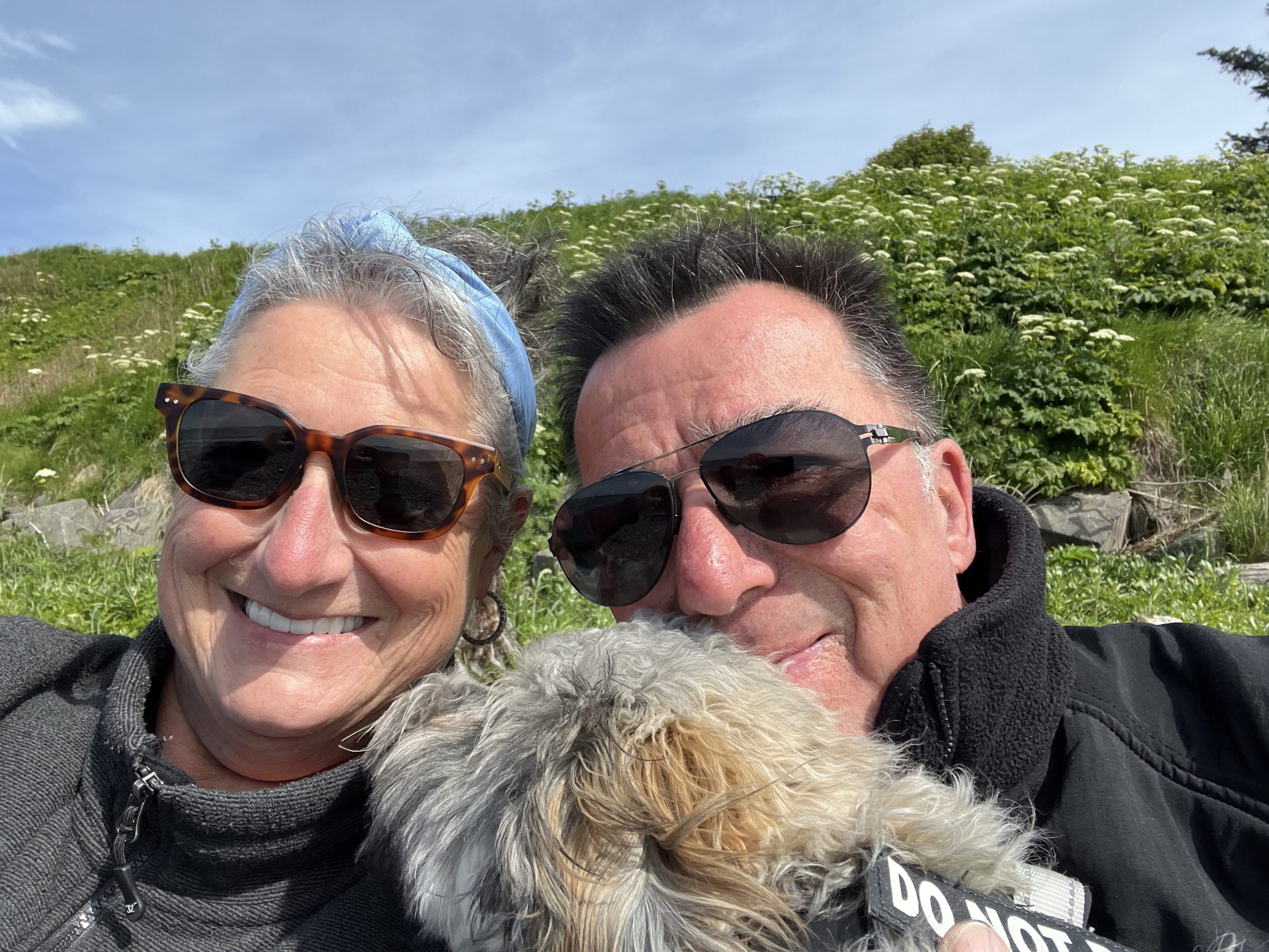 Two people wearing sunglasses smiling with a dog, green hill in background.