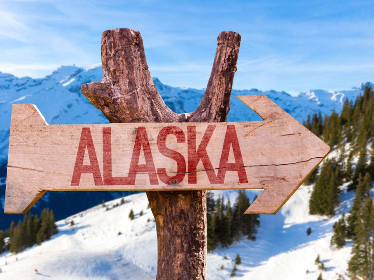 Wooden arrow sign with 'Alaska' pointing right, snowy mountains and trees in background.