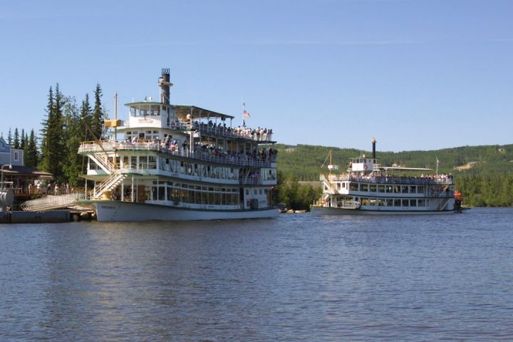 Two large riverboats with passengers sailing on a calm river, surrounded by trees and a clear sky.