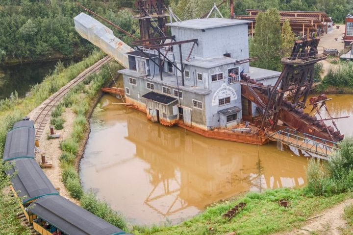 Train passing by a historic industrial dredge surrounded by trees and murky water.