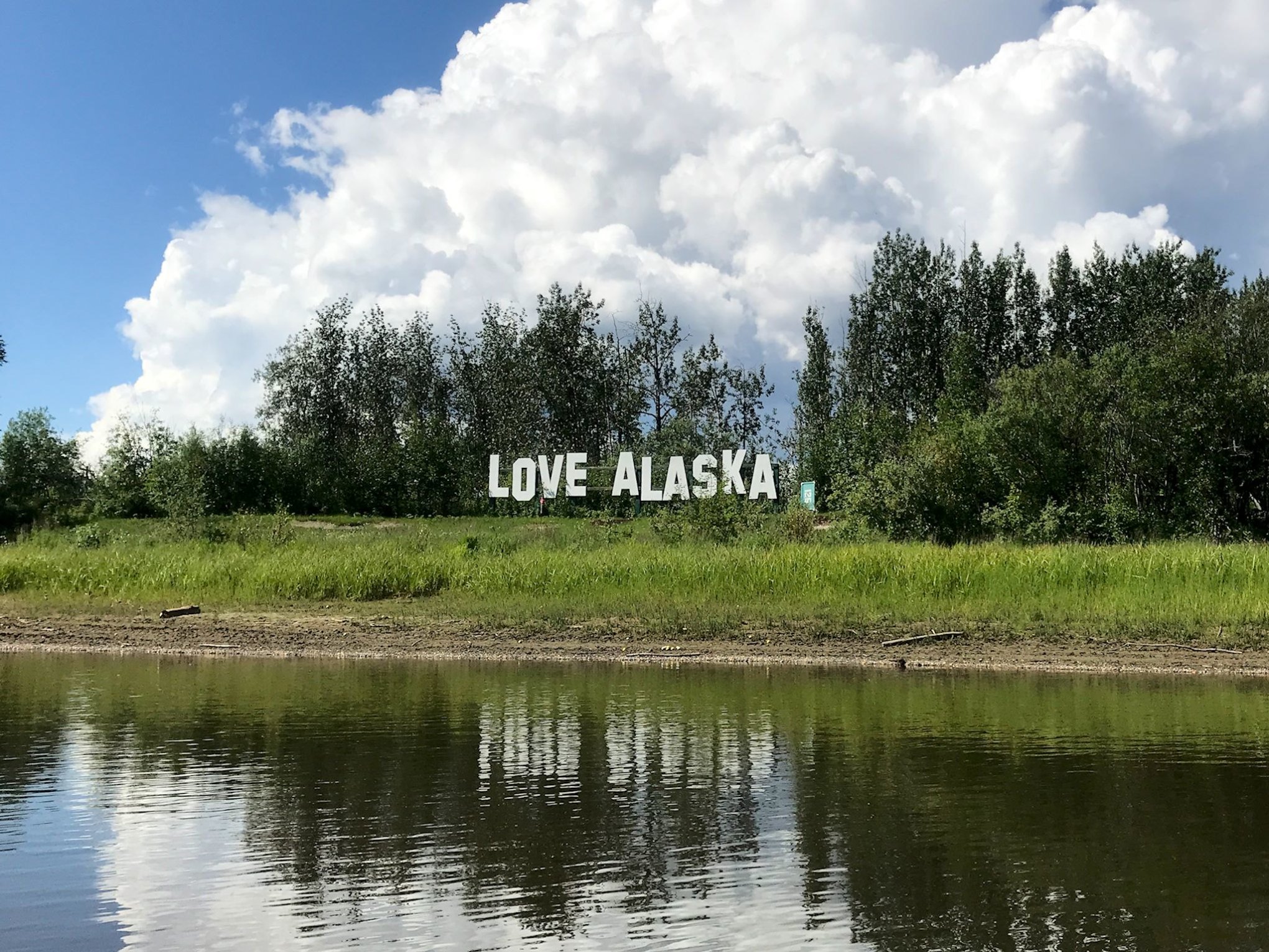 Sign reading 'LOVE ALASKA' next to trees with a river in the foreground and clouds in the sky.
