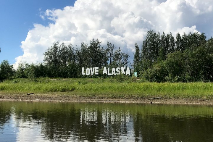 Sign reading 'LOVE ALASKA' next to trees with a river in the foreground and clouds in the sky.