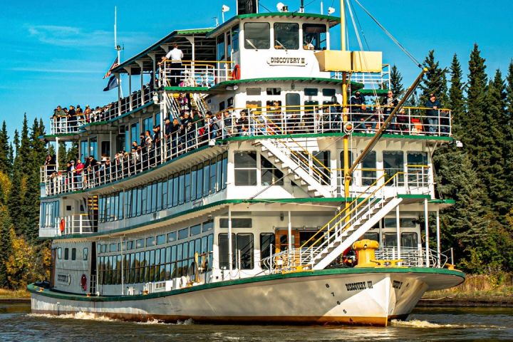 A large riverboat with multiple decks and passengers cruising on a calm river under a clear blue sky.