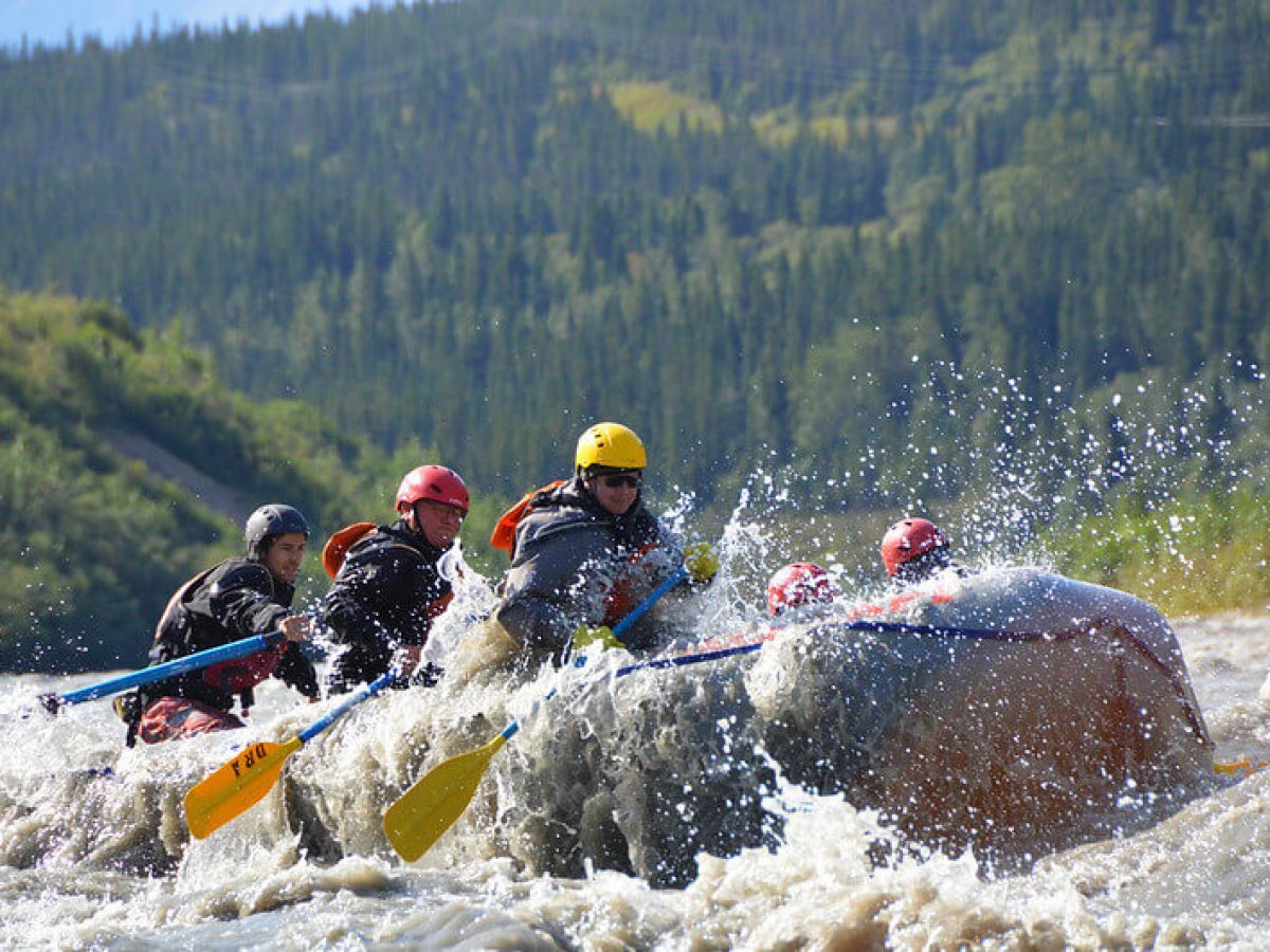 Group of people white-water rafting with paddles, surrounded by splashing water and forested hills.
