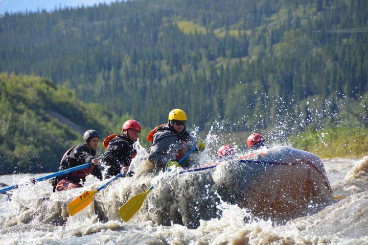 Group of people white-water rafting with paddles, surrounded by splashing water and forested hills.