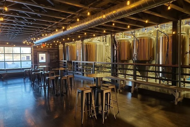 Modern brewery interior with metal tanks, wooden stools, and string lights.