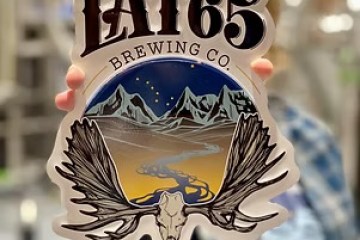 Lat65 Brewing Co. sign with moose antlers and Alaskan landscape background.