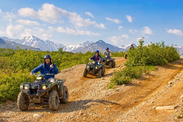 Group of people riding ATVs on a dirt trail with mountains in the background.