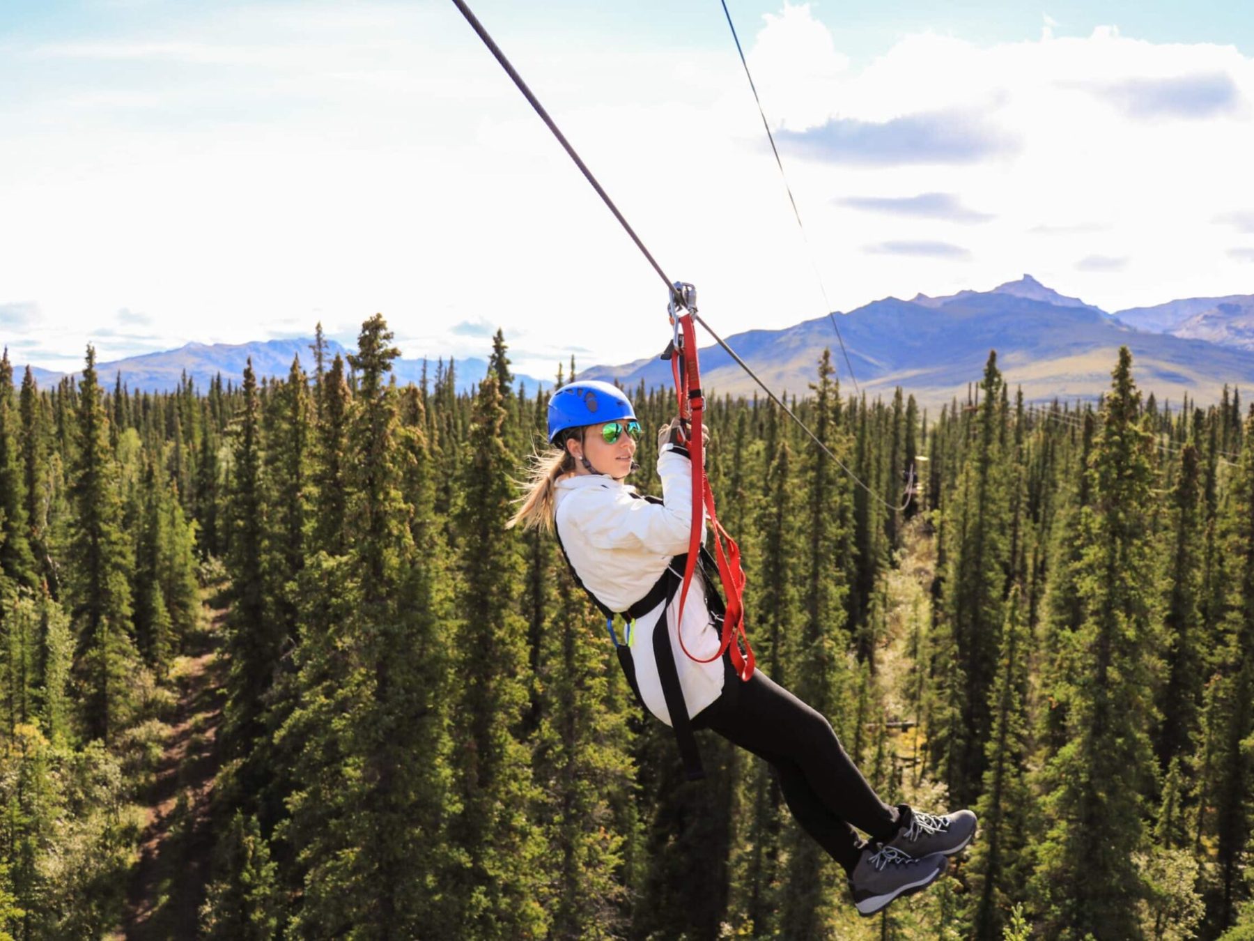 Person ziplining over a forested area with mountains in the background, wearing a helmet and harness.