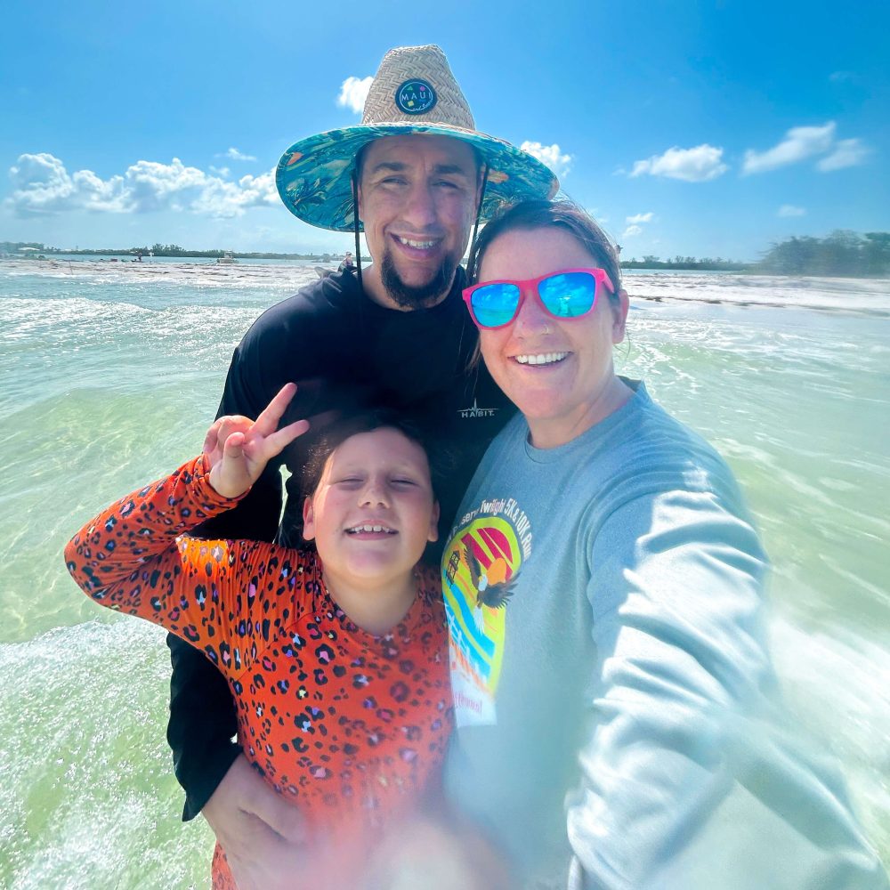 Three people smiling at the beach on a sunny day, one in a straw hat and another with sunglasses.