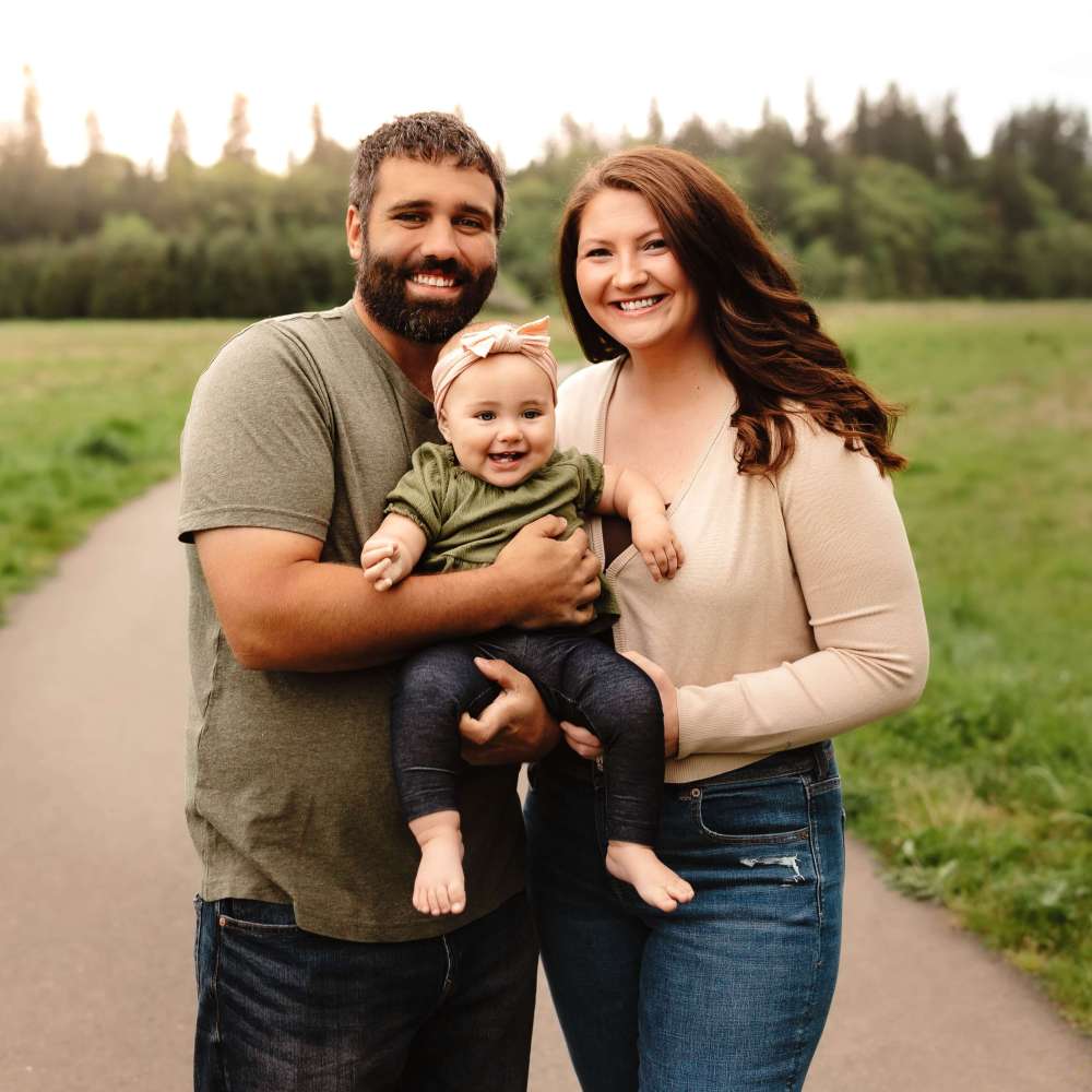 Family of three smiling on a path in a park with trees in the background.