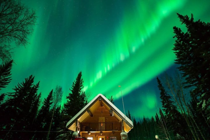 Cabin in snowy forest with bright green Northern Lights in the sky.