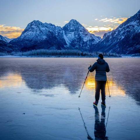 Person ice skating on frozen lake with mountains at sunset.