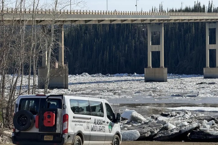 Van parked by river with ice chunks, bridge and trees in background.