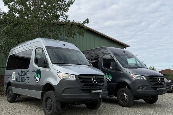 Two Mercedes vans with logos parked on gravel beside a green building.