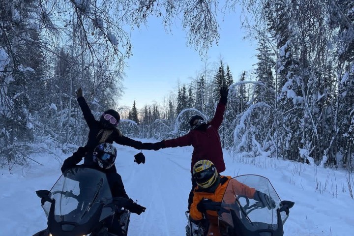 Two people on snowmobiles posing under a snow-covered tree arch.